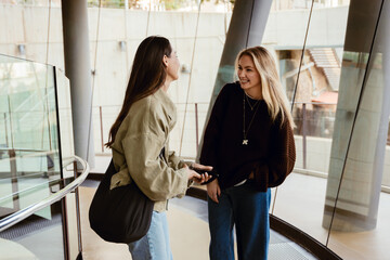 Two young White women chat happily inside a modern building with large windows and tall support...
