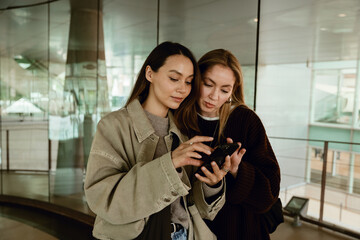 Two young White women stand side by side indoors near glass windows. They focus on a smartphone screen held by the woman on the left wearing a green jacket, viewing content.