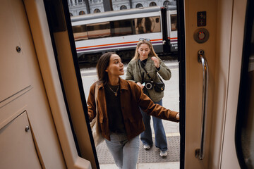 A mid-20s brunette Caucasian woman walking into the train cart while looking to the side, as her mid-20s blond Caucasian female friend follows behind her, on a train platform