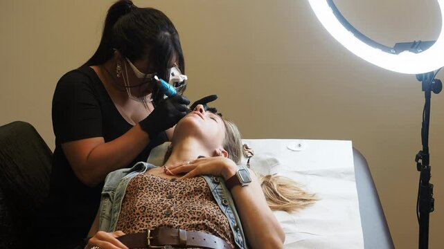 Hispanic sthetician, wearing black top and gloves, leans in carefully holding microblading tool, client lies relaxed, one hand on her chest, dressed in a leopard-print top and denim vest, slow motion.