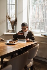 An Asian man in his 70s with dark hair, in glasses, a brown cardigan, and a white shirt, sits at a...