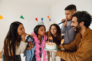 A multiethnic group of friends celebrates a birthday. A young Indian woman wearing a tiara blows...