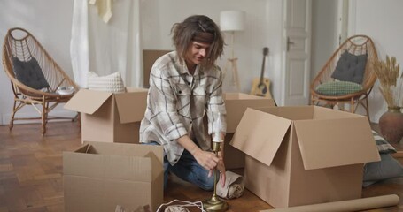 Guy disassembles lamp and puts it in cardboard box while moving house. Guy puts fragile furniture and dishes in cardboard boxes during a move