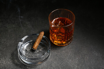 Glass of cold rum and ash tray with cigar on grey grunge background