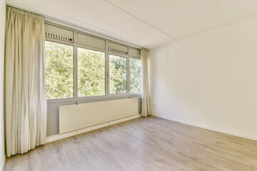 A spacious, unfurnished room featuring large windows that allow natural light to flood in, highlighting the neutral tones of the walls and wooden floor.
