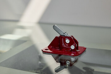 A red metal formwork latch with a silver locking mechanism placed on a reflective glass surface.