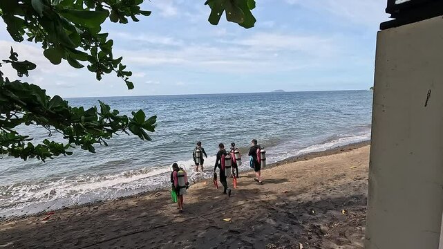 Group of divers walks on dark sandy beach to water edge preparing to enter ocean. People wear wetsuits with buoyancy compensators and diving equipment at resort