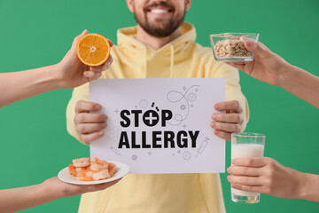 Young man with text STOP ALLERGY and female hands holding food products on green background, closeup