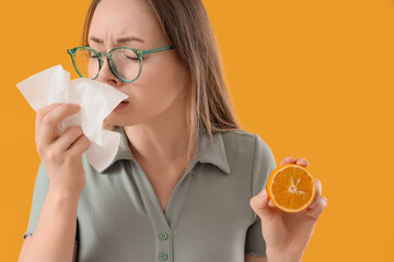 Young woman with orange and tissue sneezing on yellow background, closeup. Food Allergy Awareness...