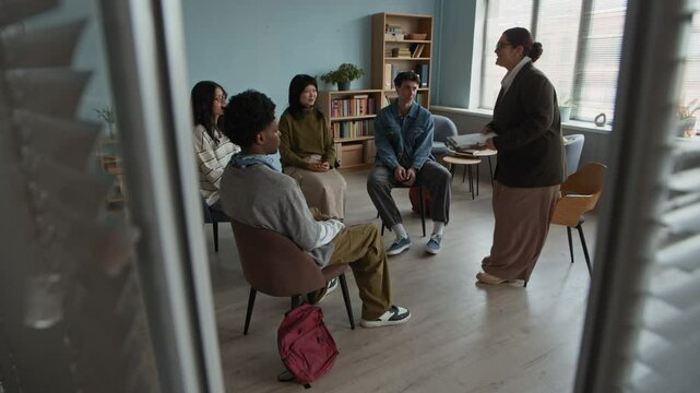 Full shot of diverse group of students sitting in circle in cozy classroom with young biracial female psychologist entering room, taking seat and introducing herself starting group therapy