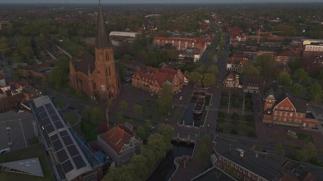 Aerial shot of Papenburg&rsquo;s city center during golden hour, featuring St. Antonius Church and the historic ship Friederike von Papenburg on the main canal. Flying backward in soft evening light.