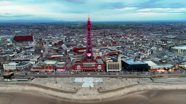 Aerial drone push-in towards the iconic Blackpool Tower, illuminated pink with a heart, overlooking the city and beach at dusk