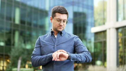 Confident businessman using smartwatch while standing on city street near modern office building. Business man with glasses looks at smart watch uses an app. Handsome male worker checks notifications - Powered by Adobe