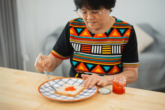 A Senior Woman Spreading Jam on Toast at a Kitchen Table, Enjoying a Moment of Quiet Breakfast with Vibrant Colors in Clothing and Tableware