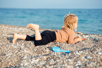 Young child girl lays and plays with sand and pebbles on beach by the sea in sunny weather