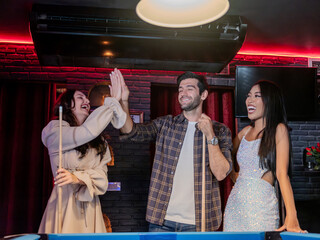 Three friends share a joyful moment at a pool table in a cozy bar. A man lines up his shot, leaning in with focus, while one woman cheerfully encourages him with a hand on his shoulder and the other w