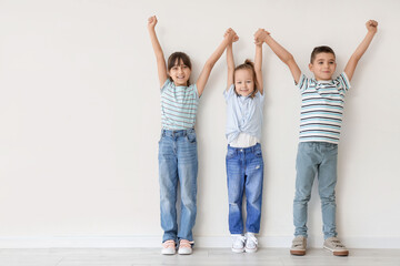 Fototapeta premium Happy diverse children holding hands with raised arms, celebrating success and friendship, standing against a white wall