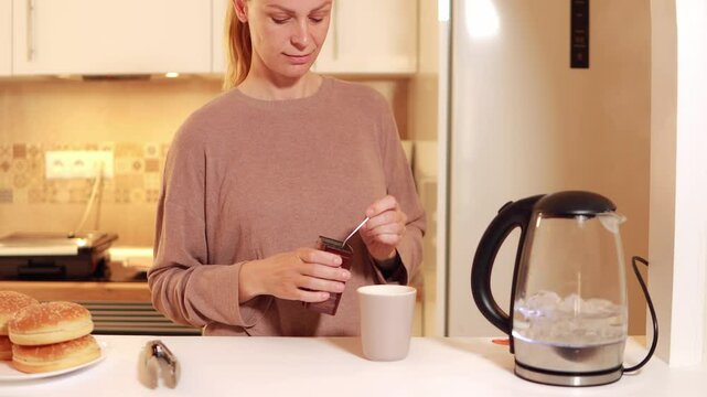 Young woman preparing instant coffee in sunlit kitchen, carefully pouring sachet contents into ceramic mug while waiting for kettle to boil, capturing morning routine moment
