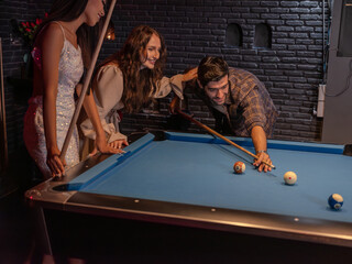 Three friends share a joyful moment at a pool table in a cozy bar. A man lines up his shot, leaning in with focus, while one woman cheerfully encourages him with a hand on his shoulder