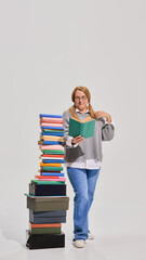 Smiling elderly woman in casual clothes standing next to high pile of books, reading with interest...