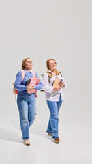 Fototapeta premium Full-length of two cheerful senior women with backpacks and books walking and talking giant white background, Elderly students. Concept of education for older adults, active aging