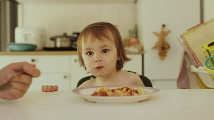 Mother feeding happy child pasta in bright kitchen at table. Joyful child happily enjoying delicious food, emphasizing importance of family bonding during mealtime