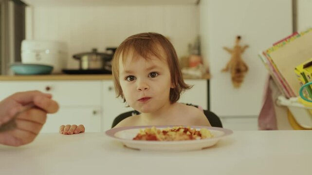 Mother feeding happy child pasta in bright kitchen at table. Joyful child happily enjoying delicious food, emphasizing importance of family bonding during mealtime