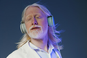 Relaxed man with long white hair enjoying music with headphones against a soft gradient background,...