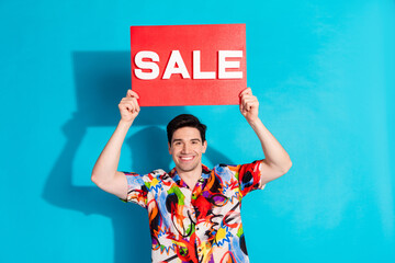 Handsome young man in colorful attire holding red sale sign against vibrant blue background,...