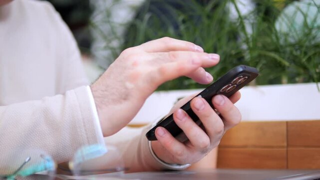 Man is using his cell phone while sitting at a table. The table is surrounded by potted plants, and there is a laptop on the table as well. Concept of relaxation and leisure
