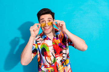 Smiling young man wearing a vibrant shirt and yellow sunglasses against a blue backdrop showing an...