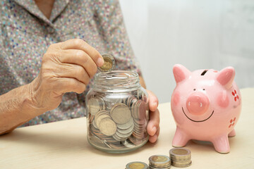 Asian elderly woman putting coin into pink piggy bank for saving money and insurance, poverty, financial problem in retirement.