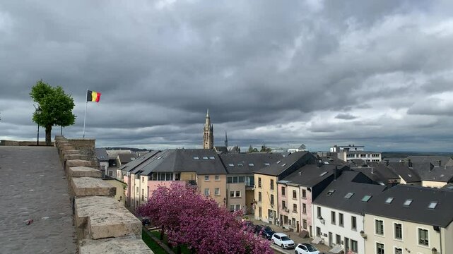 Panoramic, scenic skyline view of Arlon city buildings and Saint Martin church. Belgian flag is waving on top of Saint Donat church hill. Street with trees blooming in pink blossoms. Arlon, Belgium