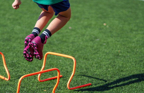 Young soccer player jumping over orange hurdles on green turf