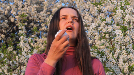 A woman is reacting to pollen allergies amidst blooming trees, expressing discomfort as she...