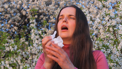 A woman stands amidst blooming trees, visibly affected by pollen allergies while holding a tissue...