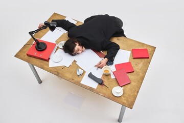 Aerial view of exhausted male employee collapsed on cluttered office desk with spilled coffee and...