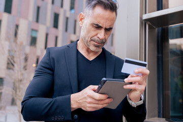 Caucasian mature man using tablet and holding a credit card to make an online payment. Focused expression in an urban environment, representing modern finance, shopping, and digital lifestyle 