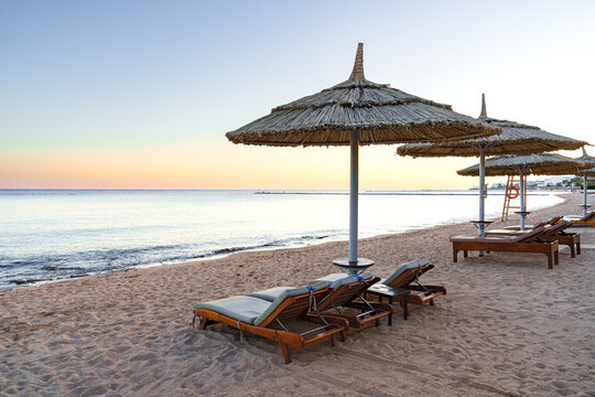 Relaxing beachside loungers under straw umbrellas at sunset near calm ocean waters
