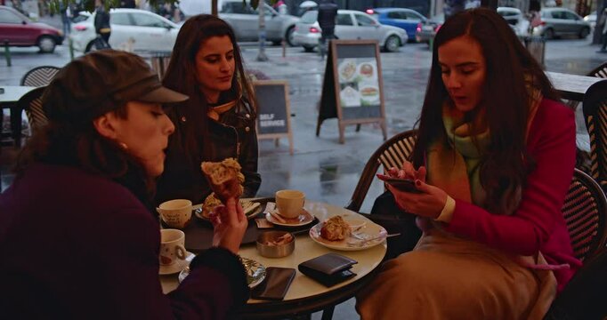 Three young women enjoy coffee and pastries at an outdoor cafe on a rainy day. They chat, check their phones, and savor the moment.