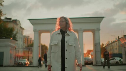 student in long white coat strolling confidently through urban square admiring sunset view as warm golden light highlights her curly hair surrounded by historic buildings people walking and cars