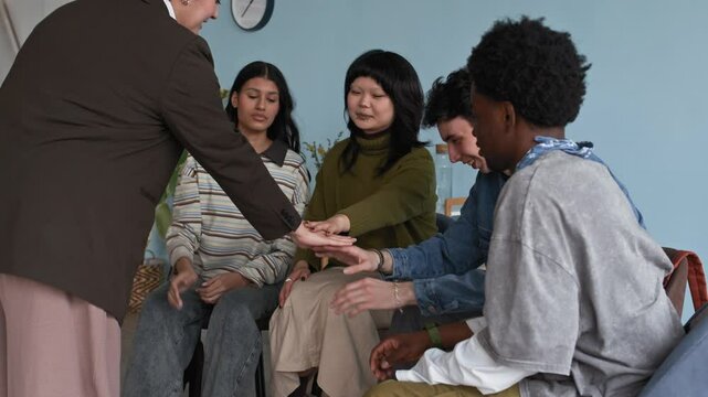 Slowmo shot of young female teacher stacking hands together with diverse teenagers sitting in circle creating positive team motivation, feeling of unity, support and cooperation