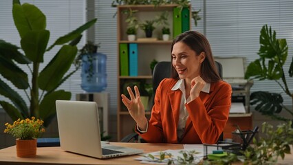 Older adult businesswoman works at green office with plants, woman sitting at desk working on laptop, talking online by video call.