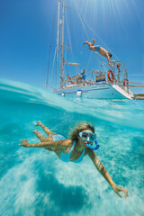 Young woman swims under water with mask, her friends are diving off a white yacht. Vertical layout