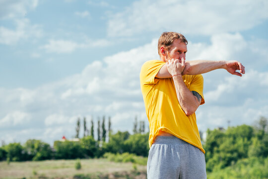 A man in a yellow t-shirt stretches his arm while standing outside on a sunny day. He is stretching his arm and shoulder muscles.