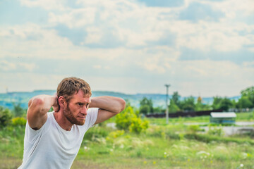 Fototapeta premium A professional sportsman is squatting in a field, with his hands behind his head. Male Performing aerobics exercises, focusing on stretching his shoulders and arms to warm up before a workout.