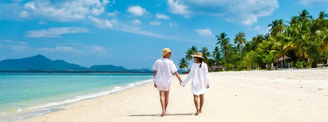 Two young Caucasian men and a Thai Asian woman walk hand in hand on the sandy shores of Koh Mook in...