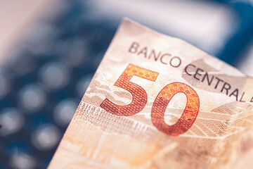 Dinheiro, Brasil, Real, 50 reais. Uma nota de 50 reais em closeup com uma calculadora desfocada ao fundo. O texto Banco Central aparecend na imagem, Destaque para o número 50. Juro alto no Brasil.