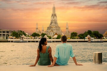 Obraz premium A couple relaxes by the Chao Praya River as they admire the stunning sunset view of Wat Arun Temple in Bangkok, Thailand. The vibrant colors of the sky create a romantic atmosphere.
