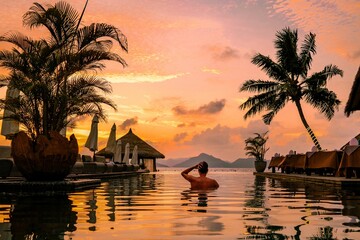 A young man enjoys a tranquil moment by a luxury swimming pool as the sun sets over the stunning...